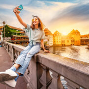 Happy and cheerful Asian girl traveler takes a selfie on the Saint Martin bridge in the Petit France area in Strasbourg. Colorful and dramatic sunset at the background